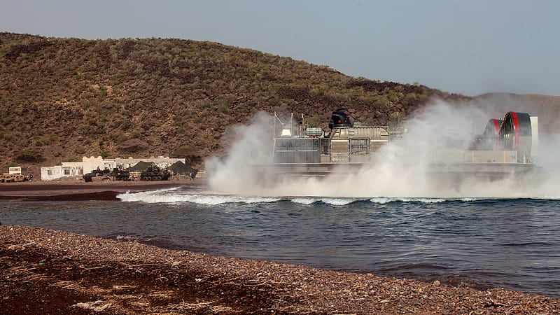 A half-submerged boat lies grounded against a muddy shoreline with its hull broken open and weathered.