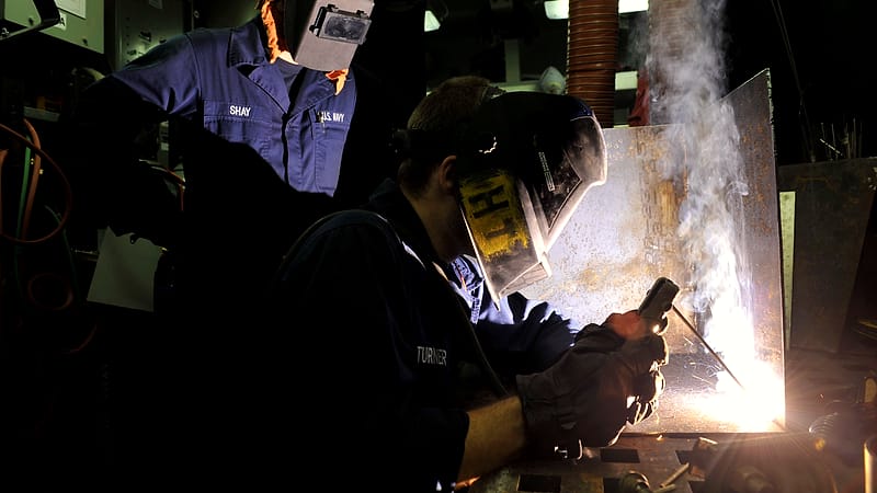 A worker in protective gear welds a metal joint in a ship's workshop while sparks fall around the bench.