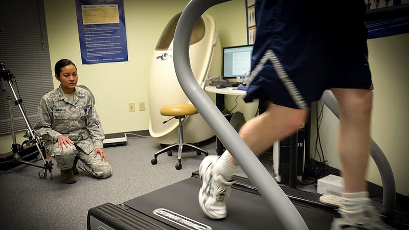 A clinician examines a patient's shoulder in a consulting room with an examination table and equipment behind them.