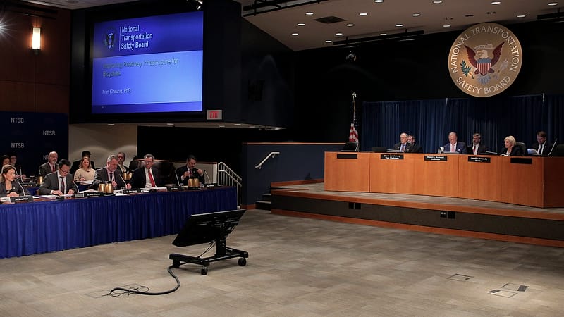 Officials seated at a curved dais with microphones and documents conduct a public board meeting before an audience.