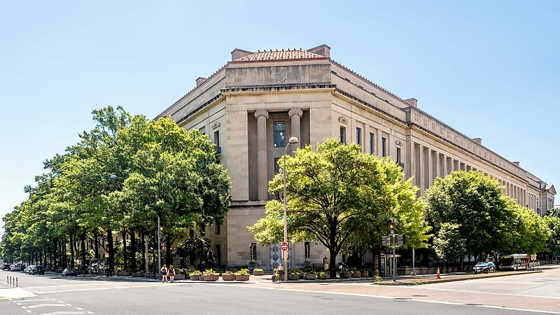 A federal government building with tall columns and flagpoles seen from across a wide plaza.
