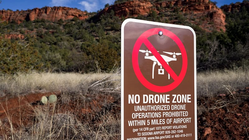 An operator holds a drone controller with both hands, watching a small quadcopter hovering in the distance over open ground.