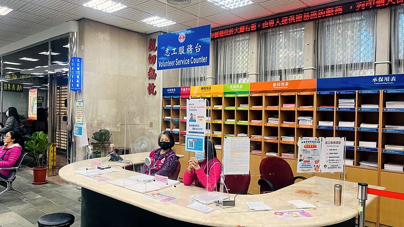 A public service counter with numbered windows, a queue barrier and a staff member seated behind glass.