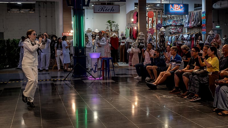 A folk ensemble in traditional dress performs on a small stage before a seated audience in a community hall.