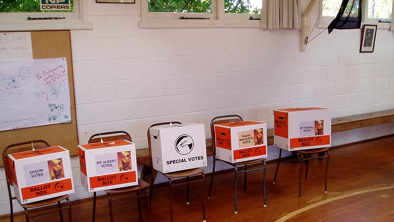 A voter places a folded ballot into a sealed box at a polling place with privacy booths behind them.