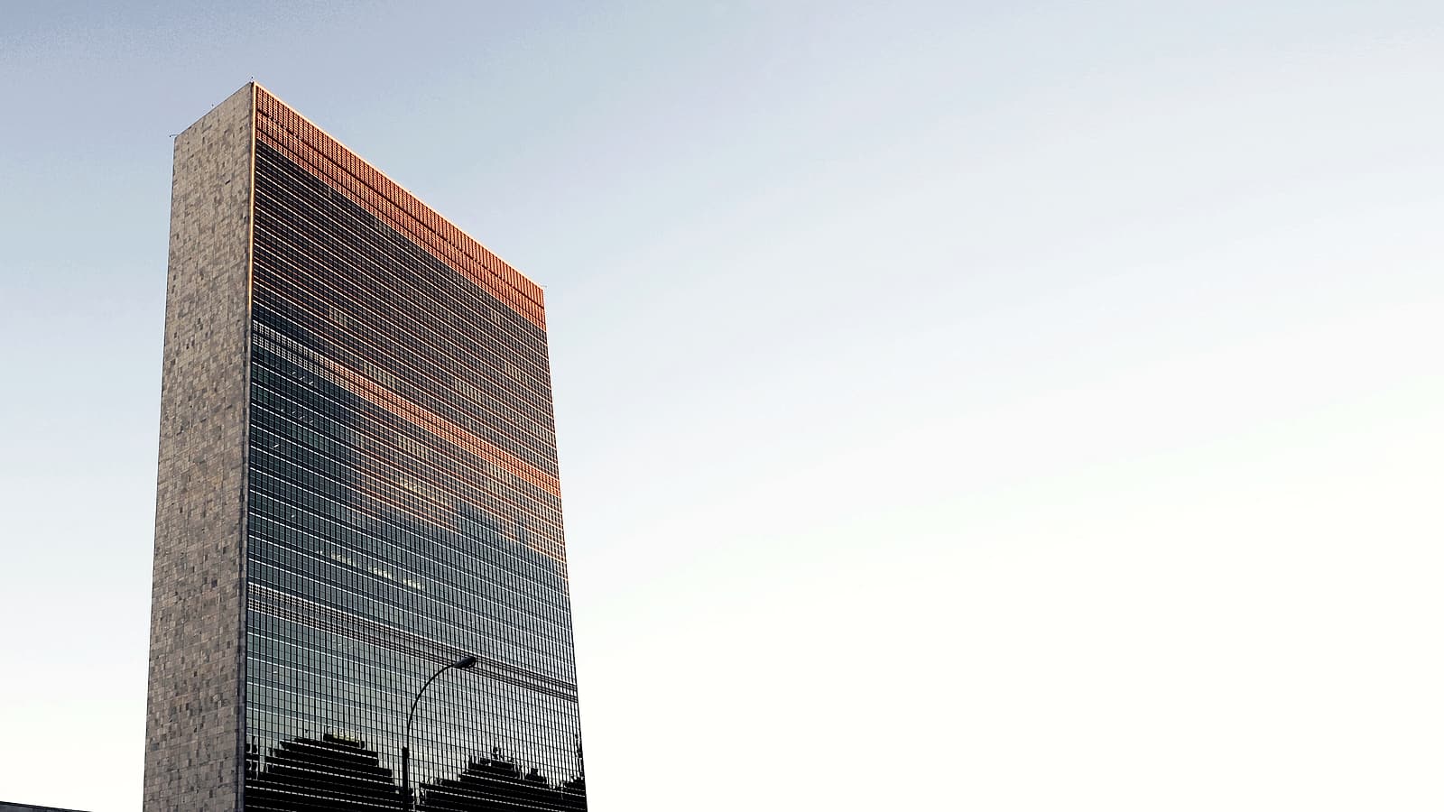 Flags of many nations stand in a row outside a modern international organization headquarters building.