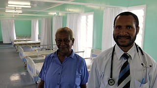 A physician and a nurse stand between beds in a hospital ward reviewing notes together.